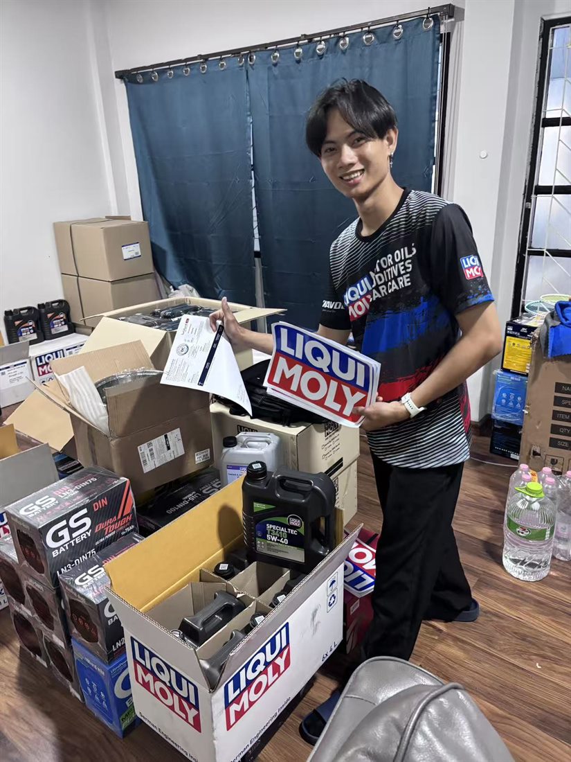 RYO พนักงานขายของ DCTHA ถือป้าย LIQUI MOLY ยืนข้างกล่องน้ำมันเครื่องและแบตเตอรี่ที่จัดเตรียมไว้ส่งลูกค้า